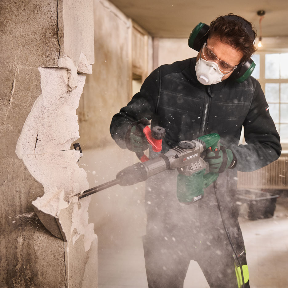 Mann mit Abbruchhammer, Maske und Gehörschutz reißt eine Wand ab.