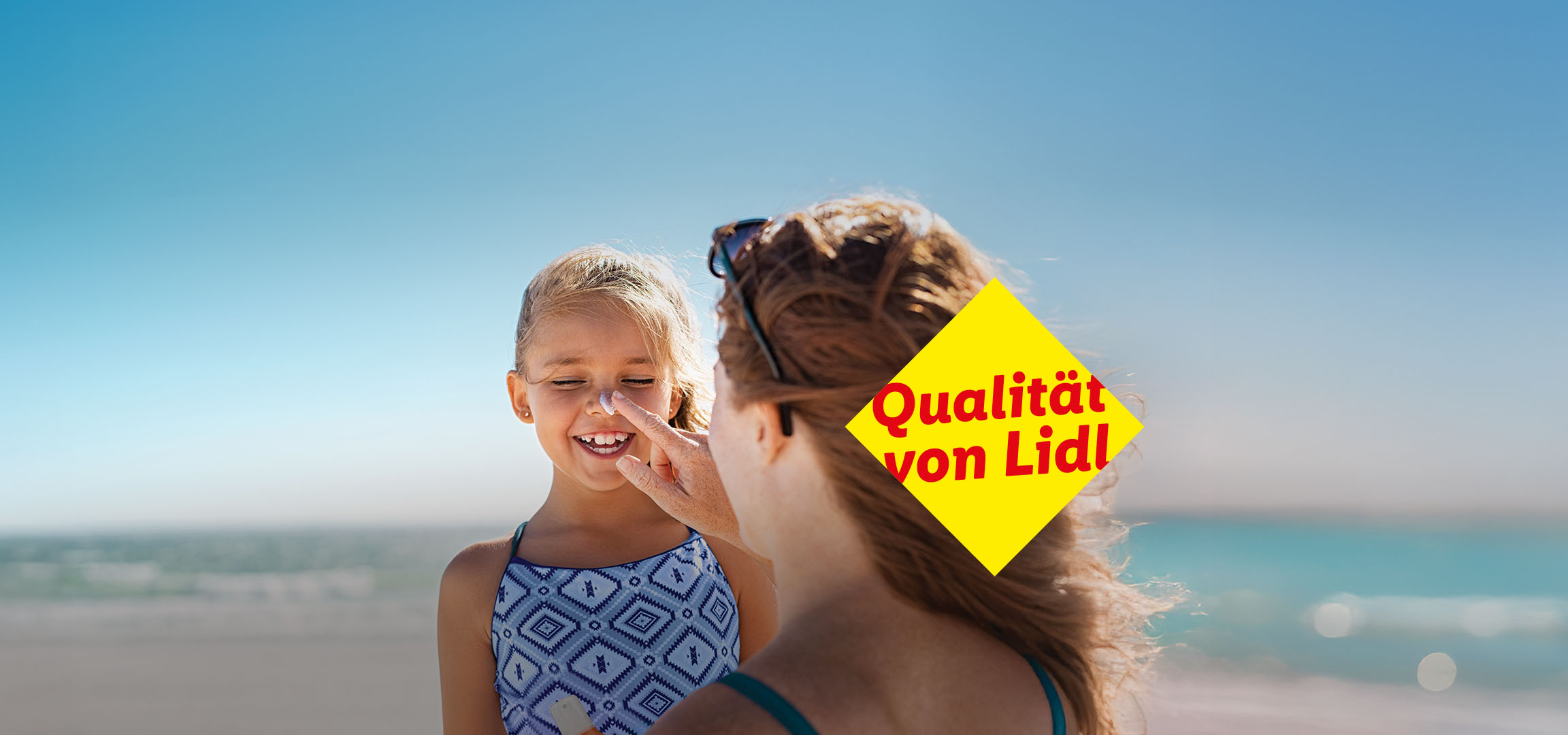 Eine Mutter cremt ihrer Tochter am Strand mit Sonnencreme ein. Textzusatz: Qualität von Lidl.