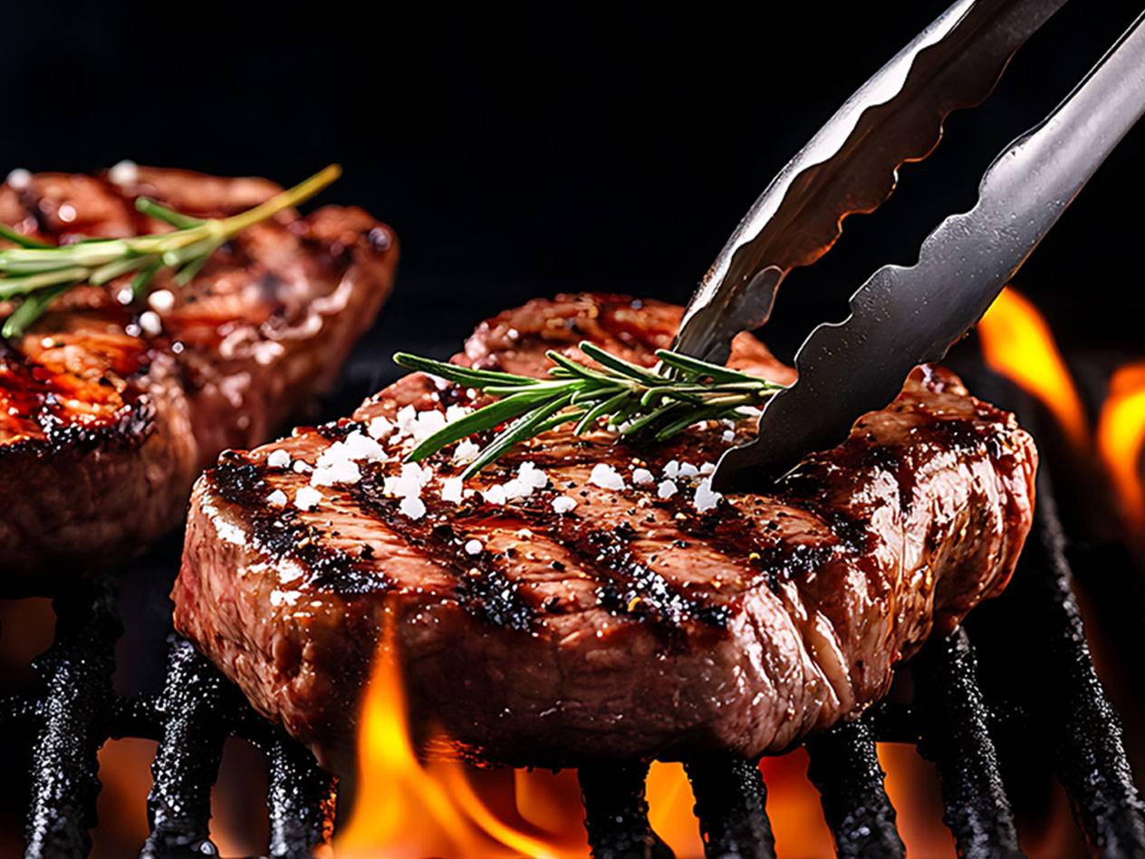 Steaks mit Rosmarin und grobem Salz auf einem Grill mit Flammen, mit einer Zange gewendet.