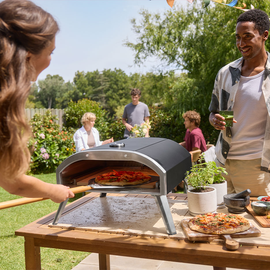 Familie auf einer Gartenparty, Frau schiebt Pizza in einen Ofen, Mann hält ein Getränk.