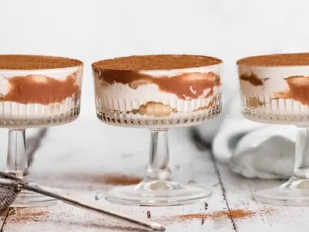 Drei Tiramisu-Desserts in Gläsern auf einem Holztisch, bestreut mit Kakaopulver.