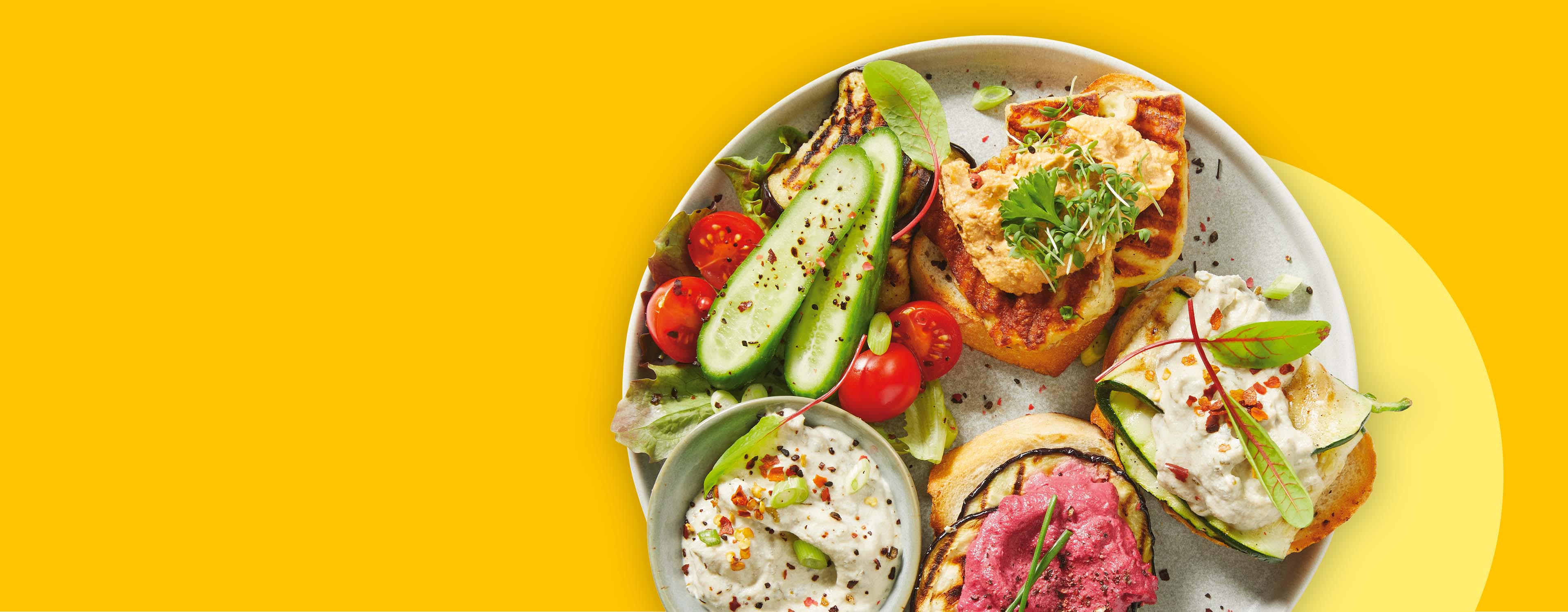 Teller mit Bruschetta, Hummus, gegrilltem Gemüse, Gurken und Tomaten auf gelbem Hintergrund.