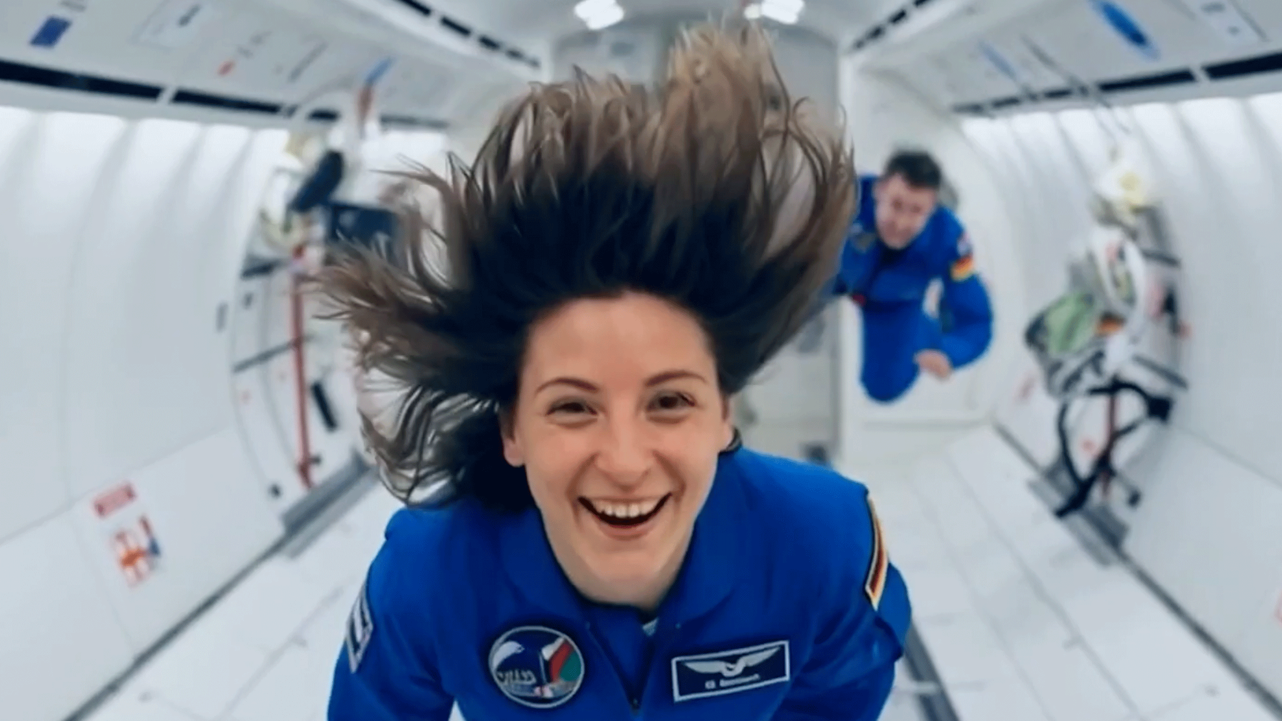 Zwei lächelnde Astronauten in blauen Overalls schweben schwerelos in einem Raumschiff.