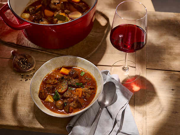 Ein Topf und eine Schale mit Rindfleischeintopf, dazu ein Glas Rotwein auf einem Holztisch.