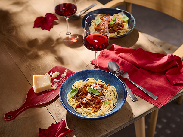 Zwei Teller Spaghetti Bolognese mit Rotwein, Parmesan und roten Servietten auf einem Holztisch.