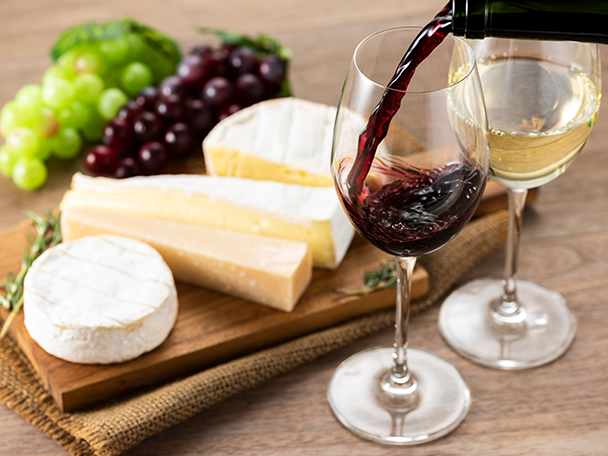 Rotwein wird in ein Glas gegossen, daneben Weißwein, Käse und Trauben auf einem Holzbrett.