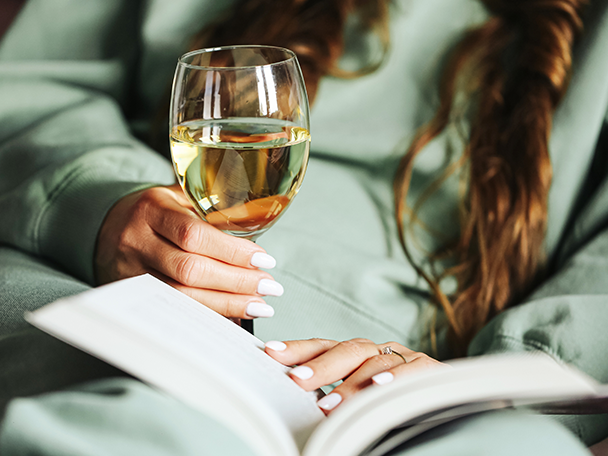 Frau mit Weißwein und Buch in der Hand, entspannt auf dem Sofa.