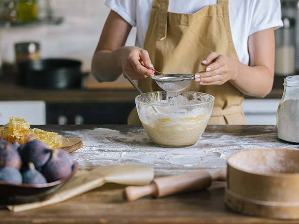 Eine Person siebt Mehl in eine Schüssel mit Teig auf einem bemehlten Holztisch.