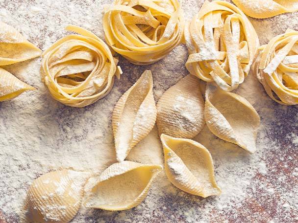 Verschiedene Nudelsorten, darunter Tagliatelle und Conchiglie, mit Mehl bestäubt auf einer Oberfläche.