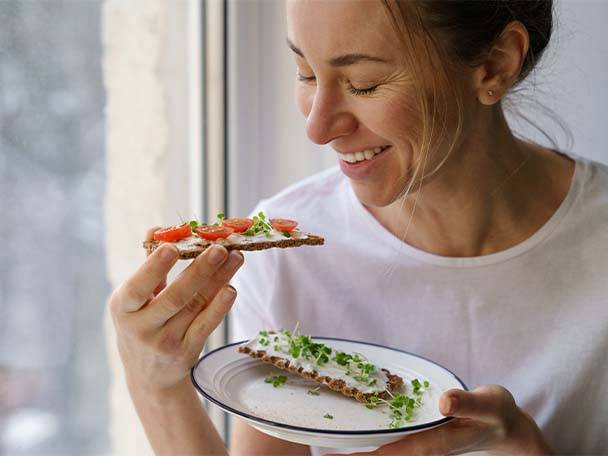 Eine lächelnde Frau hält ein Knäckebrot mit Tomaten und Sprossen, ein weiteres liegt auf einem Teller.