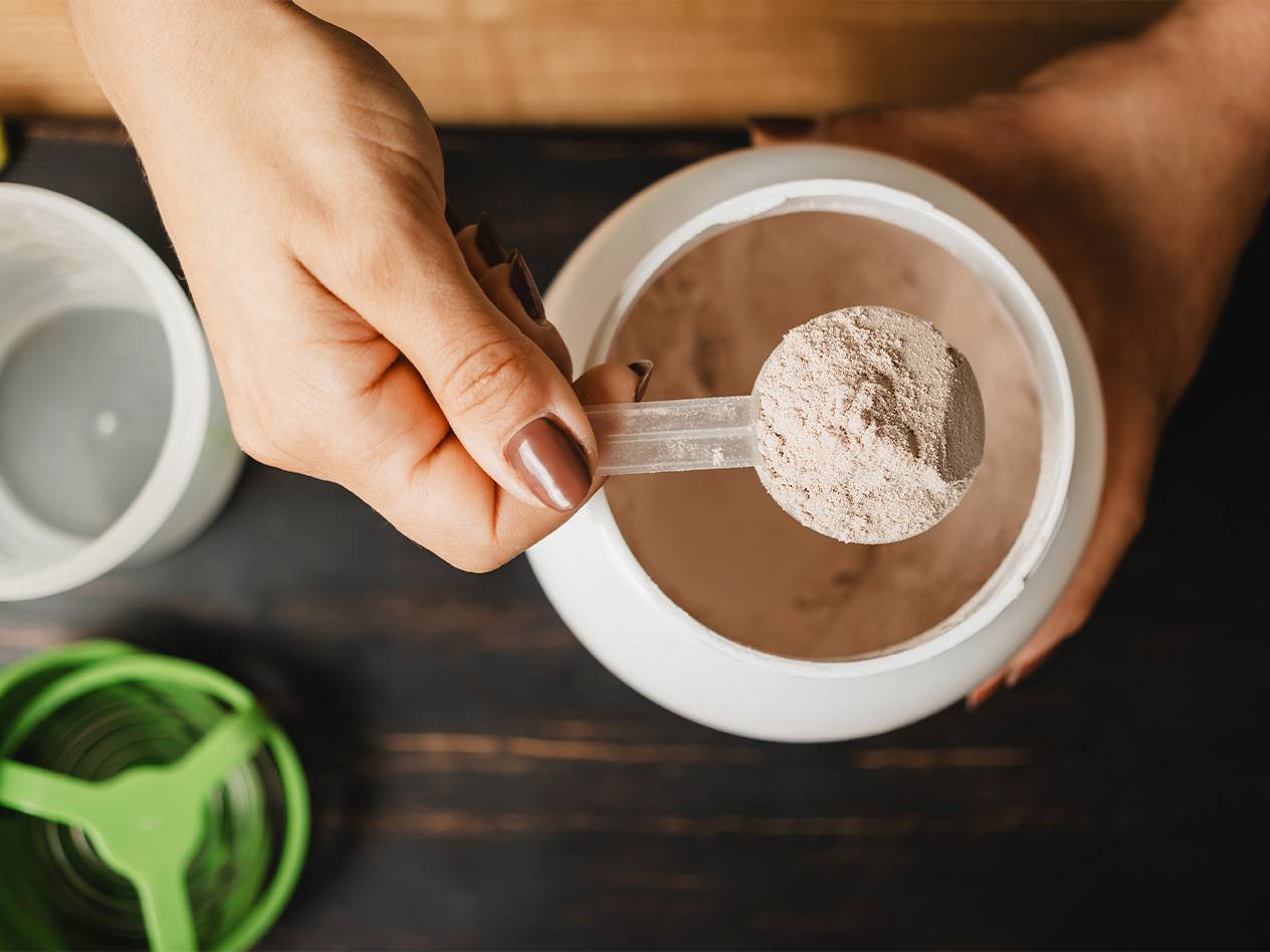 Eine Hand schöpft mit einem Messlöffel Proteinpulver aus einem weißen Behälter.