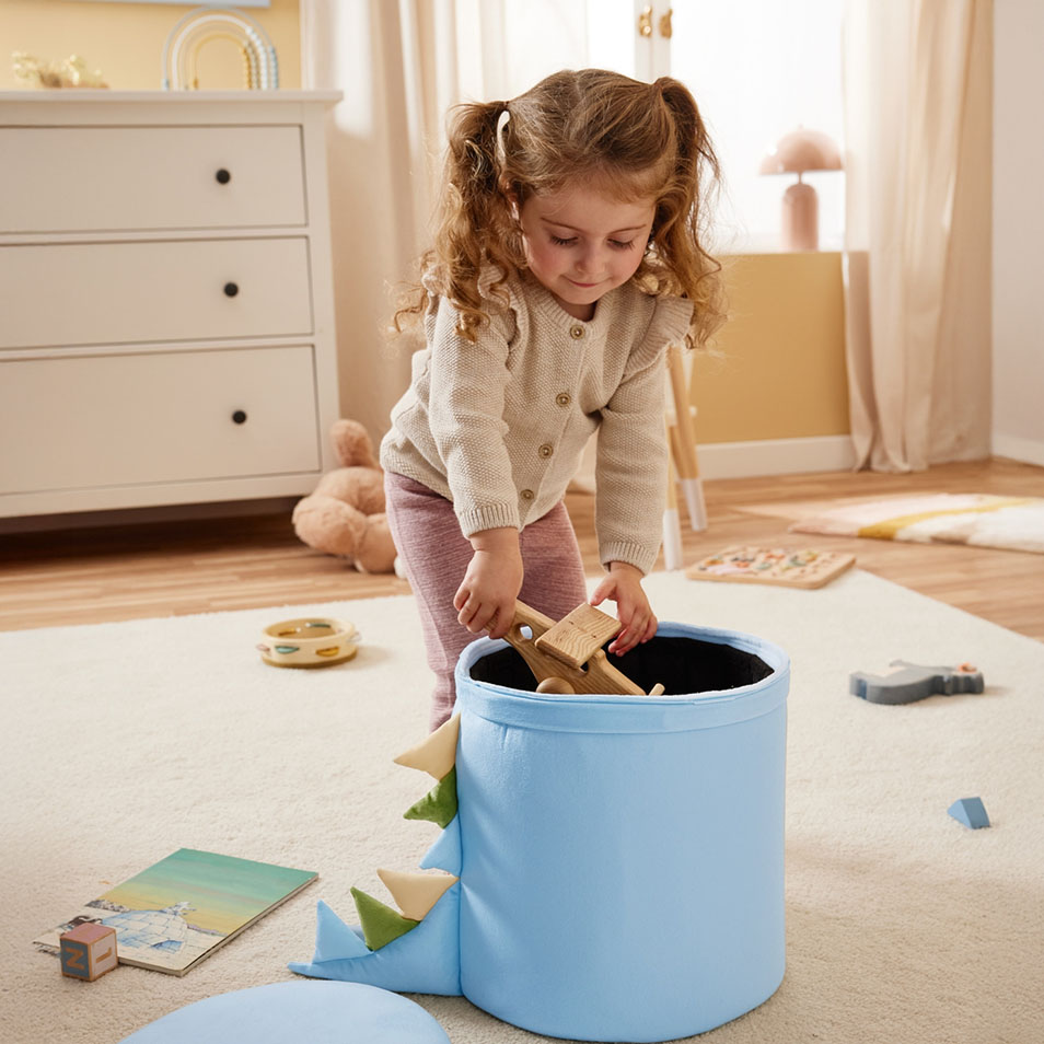 Ein Kind legt Holzspielzeug in eine blaue Aufbewahrungsbox mit Dinosaurier-Motiv in einem Schlafzimmer.