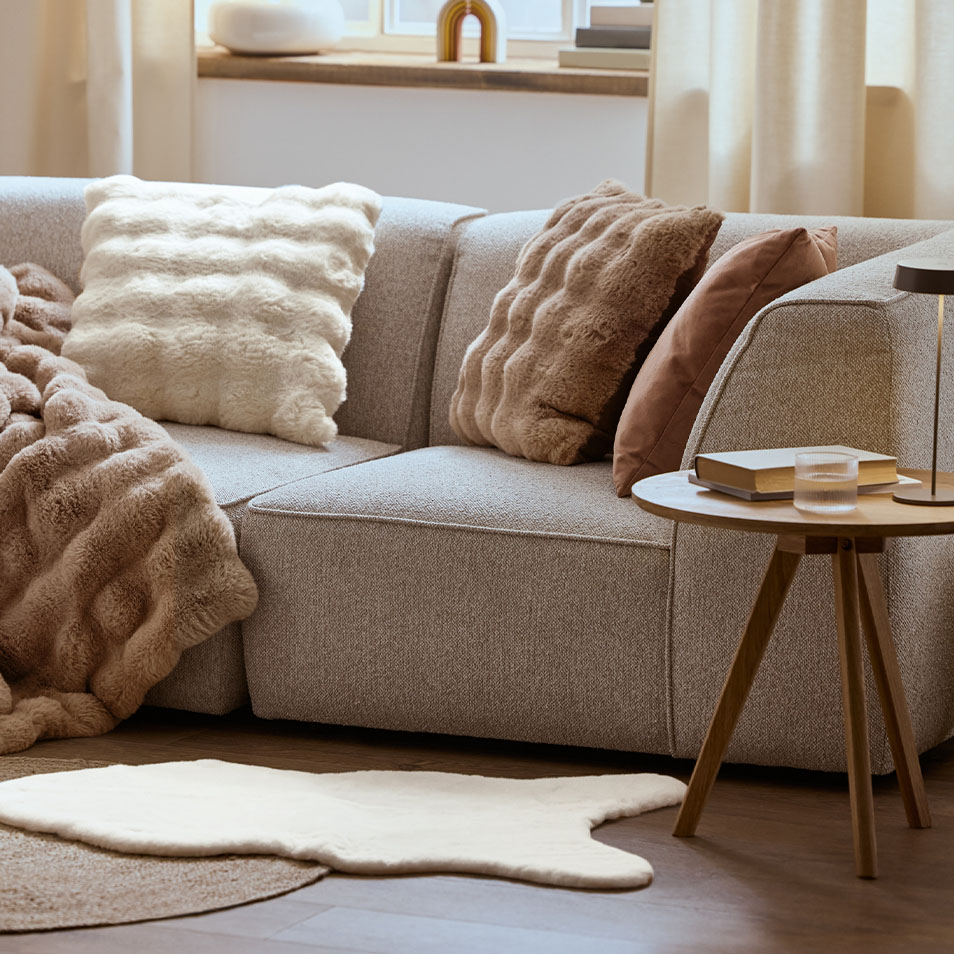 Ein graues Modulsofa mit flauschigen Kissen und einer Decke, neben einem Beistelltisch aus Holz.